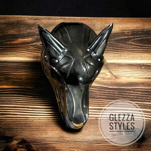 May include: A black, stylized Anubis mask with pointed ears and a metallic finish. The mask is set against a wooden background and features the text "GLEZZA STYLES" in a circular logo.