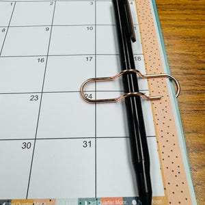 May include: A close-up of a 2025 calendar with a black pen and a gold paper clip. The calendar has a colorful design with a light blue background and a white grid.