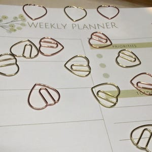 May include: A collection of heart-shaped paper clips in gold and rose gold tones scattered on a weekly planner. The planner has the words "WEEKLY PLANNER" printed on it.