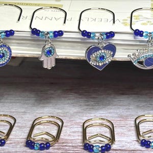 May include: A collection of decorative paper clips with charms. The charms include an evil eye, a Hamsa hand, an eye in a heart, and a crown with an eye. The clips are gold-toned with blue and turquoise beads.