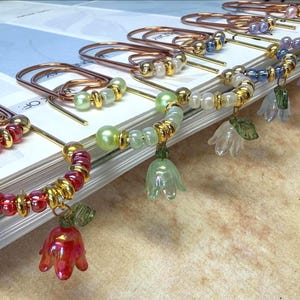 May include: A collection of large copper-toned metal paperclips, each adorned with a decorative charm featuring a bell-shaped flower bead in various colors, accented by smaller round metallic gold beads and translucent glass beads in matching hues.
