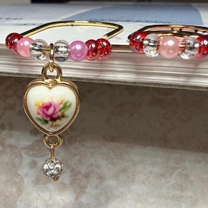 May include: Two gold-toned rings featuring a series of red, pink, and clear beads. The left ring includes a heart-shaped charm with a floral rose design and a small dangling crystal bead. The right ring is a simple band adorned with similar beads.