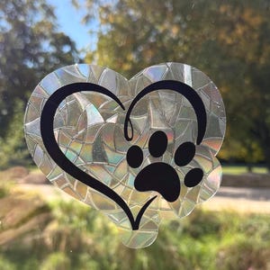 May include: A heart-shaped window cling with a black paw print and heart design. The cling has a holographic, iridescent finish, reflecting light. The background shows a blurred view of trees and sunlight.