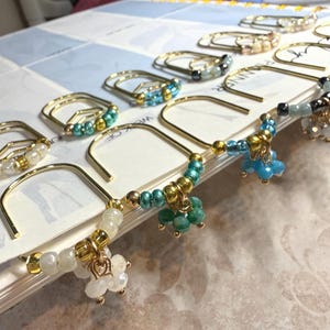 May include: A collection of gold-toned metal paper clips, each embellished with a unique arrangement of colorful glass beads and small dangling charms. The beads feature various shades of blue, green, white, and metallic gold, creating a decorative set.