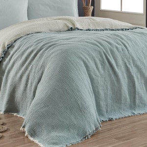 Puede incluir: Una manta de tiro azul claro y blanco con borde de flecos. La manta está doblada en una cama con dos fundas de almohada azul claro.