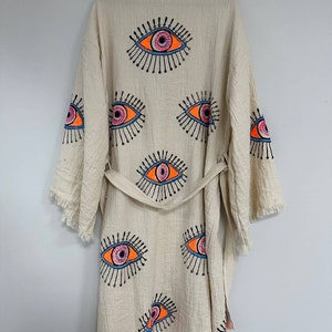 May include: A cream-colored robe with a fringe trim and a tie belt. The robe is decorated with multiple colorful eye designs.