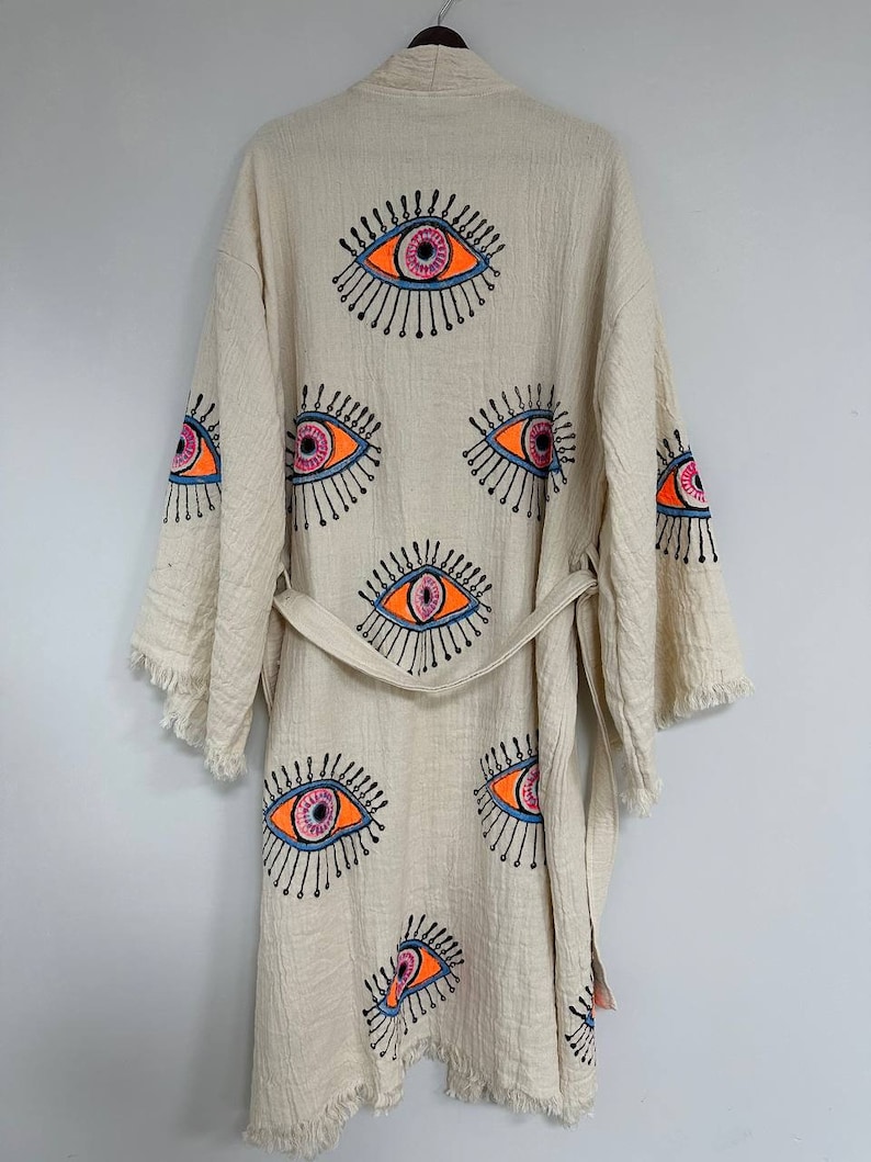 May include: A cream-colored robe with a fringe trim and a tie belt. The robe is decorated with multiple colorful eye designs.