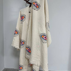 May include: A white cotton robe with a fringe trim and a pattern of colorful eyes. The robe is hanging on a wooden stand.