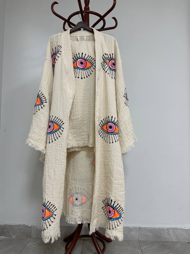May include: A white cotton robe with a long, flowing design. The robe features a colorful eye pattern throughout. The robe is on a wooden hanger.