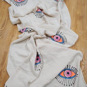 May include: A white cotton throw blanket with a fringed edge features a repeating pattern of colorful evil eyes. The eyes are outlined in black and filled with pink, blue, and orange.