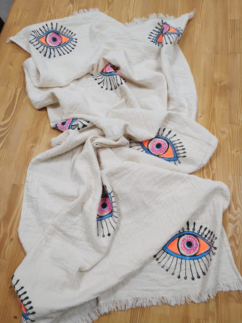 May include: A white cotton throw blanket with a fringed edge features a repeating pattern of colorful evil eyes. The eyes are outlined in black and filled with pink, blue, and orange.