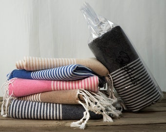 Könnte beinhalten: Ein Stapel gestreifter Handtücher in verschiedenen Farben, darunter rosa, blau, beige und schwarz. Die Handtücher sind gefaltet und haben weiße Fransen. Das schwarze Handtuch befindet sich in einer durchsichtigen Plastiktüte.