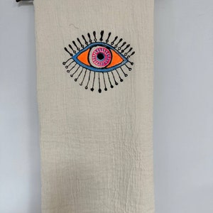 May include: A white cotton throw blanket with a black, blue, orange, and pink evil eye design.