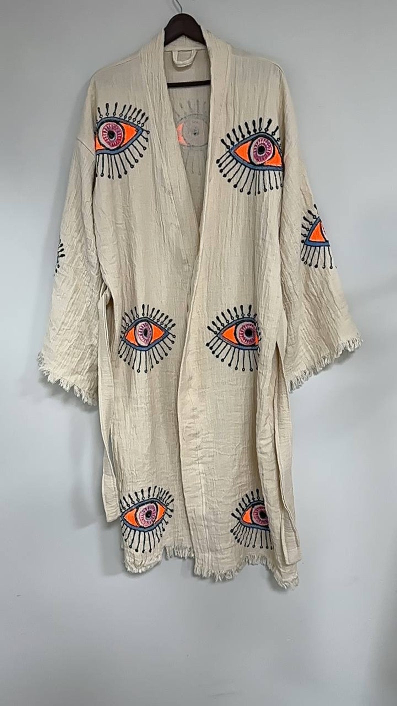 May include: A cream-colored robe with a fringed hem and a tie belt. The robe is decorated with embroidered eyes in shades of orange, pink, blue, and black.