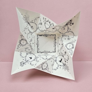 Peut inclure: Un papier blanc de diseuse de bonne aventure, plié en forme d'étoile, avec des illustrations florales et botaniques noires. Le motif comprend un carré central et des cadres circulaires avec du texte. Le fond est rose uni.