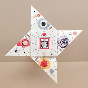 Peut inclure: Une étoile en papier plié avec un carré central portant le mot "ARIES" dans un cadre rouge. Les pointes de l'étoile présentent des illustrations célestes avec des planètes, des étoiles et des symboles du zodiaque. La palette de couleurs comprend le rouge, le bleu, le jaune et le blanc.