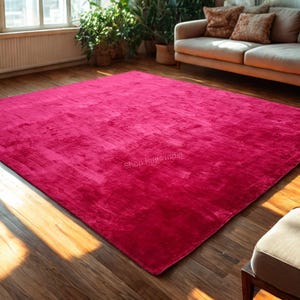May include: A large, rectangular magenta rug dominates a room with hardwood floors. The rug has a textured appearance and is placed in front of a beige sofa and a window. The text "Shop Rugs India" is visible on the rug.