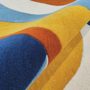 May include: A tufted area rug featuring a bold, curved pattern with distinct bands of mustard yellow, deep navy blue, burnt orange, light blue, and off-white. The rug is folded over, revealing the thick, textured pile and the layered color design.