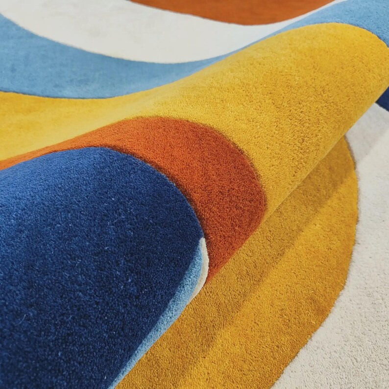 May include: A tufted area rug featuring a bold, curved pattern with distinct bands of mustard yellow, deep navy blue, burnt orange, light blue, and off-white. The rug is folded over, revealing the thick, textured pile and the layered color design.