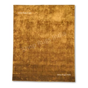 May include: A rectangular, golden-brown rug with a textured appearance. The rug has a mottled pattern with darker brown areas. The words "Shop Rugs India" are printed on the rug in multiple places.