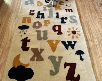 Könnte beinhalten: Ein cremefarbener Teppich mit dem Alphabet in bunten Buchstaben gedruckt. Die Buchstaben sind in einer verspielten, fantasievollen Schriftart. Der Teppich zeigt auch ein paar einfache Illustrationen, darunter eine Wolke, eine Sonne, einen Mond und einen Regenschirm.