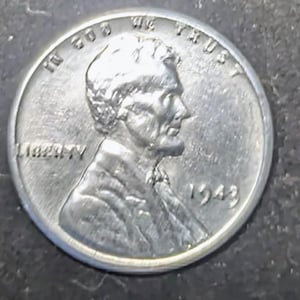 May include: A close-up of a silver 1943 Lincoln penny. The coin shows the profile of Abraham Lincoln, the word "LIBERTY", and the phrase "IN GOD WE TRUST". The year "1943" is visible.