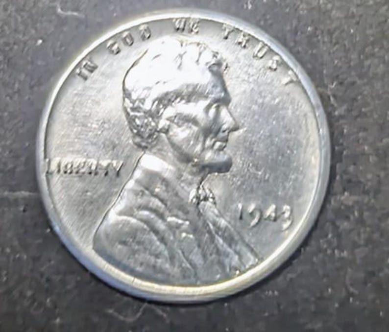 May include: A close-up of a silver 1943 Lincoln penny. The coin shows the profile of Abraham Lincoln, the word "LIBERTY", and the phrase "IN GOD WE TRUST". The year "1943" is visible.