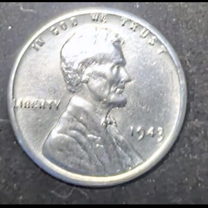 May include: A close-up of a silver 1943 Lincoln penny. The coin features a profile of Abraham Lincoln, with the words "LIBERTY" and "IN GOD WE TRUST" visible. The year "1943" is also displayed.