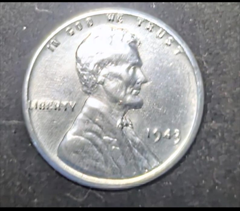 May include: A close-up of a silver 1943 Lincoln penny. The coin features a profile of Abraham Lincoln, with the words "LIBERTY" and "IN GOD WE TRUST" visible. The year "1943" is also displayed.