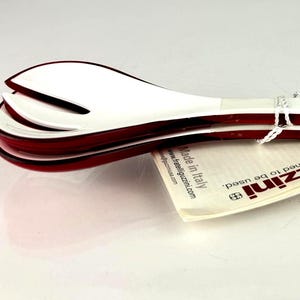 Puede incluir: Un juego de cubiertos para ensalada blancos y burdeos, con un diseño de cuchara y tenedor ranurados. Los cubiertos tienen una estética elegante y moderna y están atados con una cuerda blanca. La etiqueta dice "guzzini designed to be used".