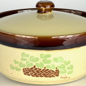 Puede incluir: Una fuente de cerámica vintage con tapa. La fuente es de color crema con borde y asas marrones. Un diseño de cesta y hojas verdes y marrones en el lateral. La tapa tiene un pomo marrón.