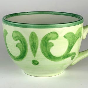 May include: A ceramic mug with a pale cream base color featuring hand-painted green scrollwork patterns around the exterior. The mug has a wide, rounded bowl shape, a single curved handle on the right side, and a solid green rim along the top edge.