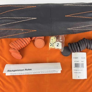 May include: A backgammon set with orange and brown game pieces, dice, and a rule sheet. The set includes a black leather case with orange and white stitching, and an orange velvet carrying bag. The label reads "Perry Ellis".