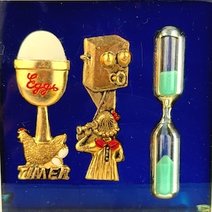 Puede incluir: Una pieza decorativa cuadrada azul con tres imágenes en relieve doradas. Las imágenes incluyen un reloj de huevos con la palabra "Eggs", una niña con un telescopio y un reloj de arena. También es visible la palabra "TIMER".