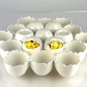May include: A white ceramic egg holder with a scalloped edge design. The holder contains several egg-shaped indentations, with two ceramic chicks emerging from cracked eggshells. The chicks are yellow with brown and red accents.