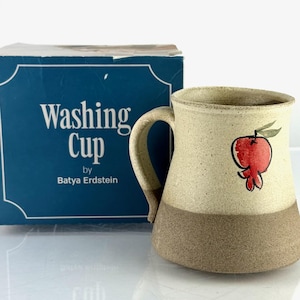 Op de afbeelding: Een keramische wasbeker van Batya Erdstein met een rood granaatappelontwerp. De mok heeft een beige bovenste helft en een bruine onderste helft. De mok staat naast een blauwe doos met de woorden "Washing Cup" erop.