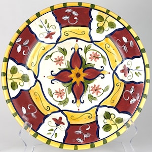 May include: A round ceramic plate featuring a central yellow flower motif surrounded by red and green floral patterns. The design includes alternating panels of deep red and white, with a yellow border accented by dark green vertical stripes.