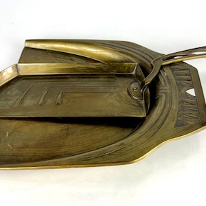 Puede incluir: Un juego antiguo de recogedor y cepillo de latón. El recogedor tiene un diseño curvo con un asa, y el cepillo encaja perfectamente en su interior. El conjunto presenta patrones geométricos de estilo Art Déco y está hecho de latón.