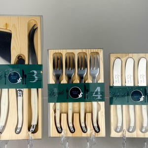 Pode incluir: Três conjuntos de facas de queijo e utensílios de servir Laguiole, quatro garfos e quatro facas de manteiga. Cada conjunto é apresentado numa caixa de madeira com uma tampa de plástico transparente. As pegas são creme e as lâminas são prateadas.