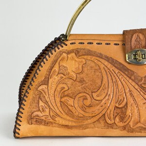May include: A tan leather handbag with a floral embossed design. The bag features a brass handle and a clasp closure. The edges are stitched with dark brown thread, and the bag has a vintage aesthetic.