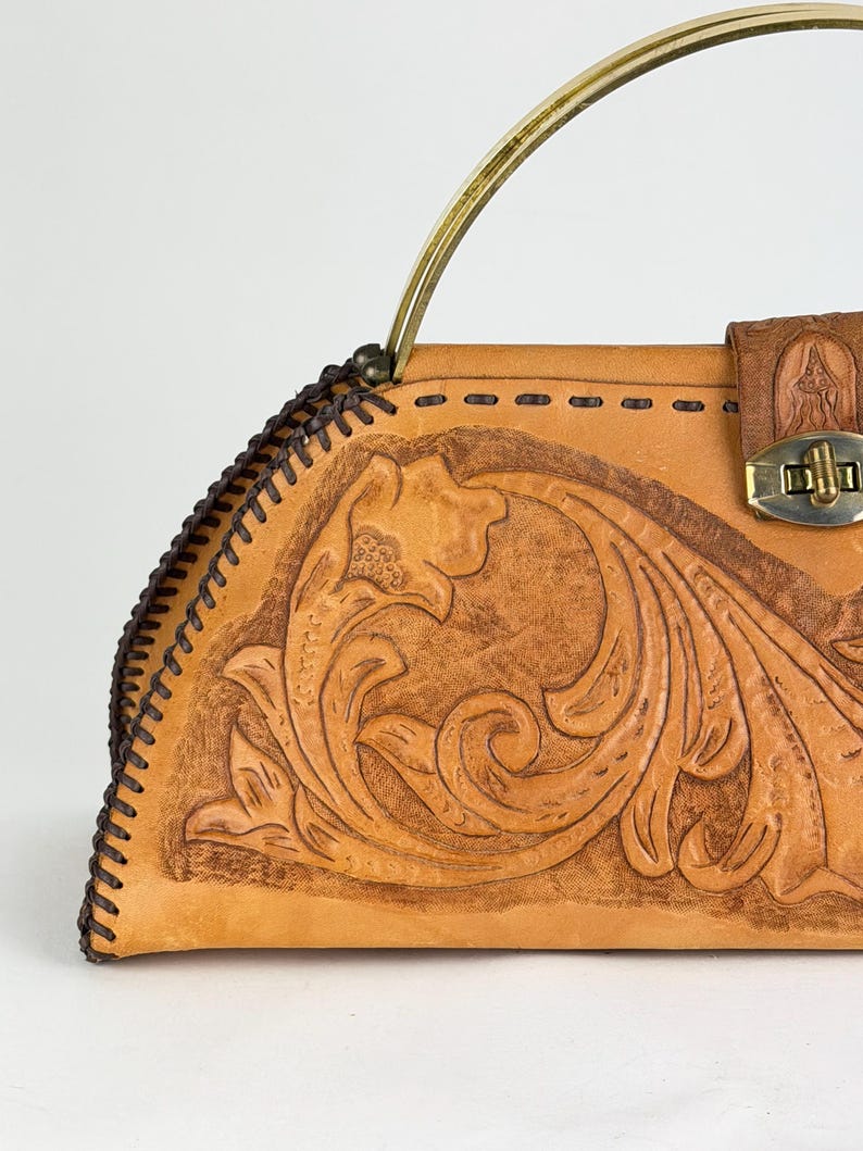 May include: A tan leather handbag with a floral embossed design. The bag features a brass handle and a clasp closure. The edges are stitched with dark brown thread, and the bag has a vintage aesthetic.