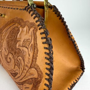 May include: A tan leather handbag with a floral embossed design. The bag features a braided brown trim and brass handles. The interior is a lighter tan color. The bag is open, revealing the inside.