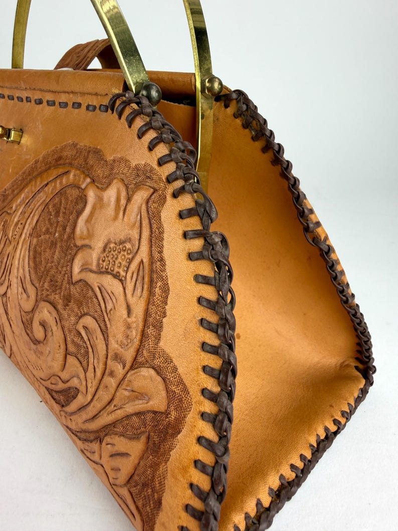 May include: A tan leather handbag with a floral embossed design. The bag features a braided brown trim and brass handles. The interior is a lighter tan color. The bag is open, revealing the inside.