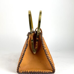 May include: A vintage tan leather handbag with a trapezoid shape. The bag features a brass frame and handles, with dark brown stitching along the edges. The interior is visible, and the bag is set against a white background.