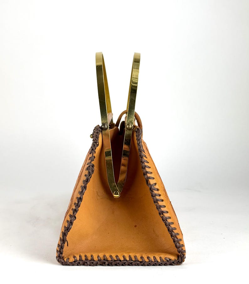 May include: A vintage tan leather handbag with a trapezoid shape. The bag features a brass frame and handles, with dark brown stitching along the edges. The interior is visible, and the bag is set against a white background.
