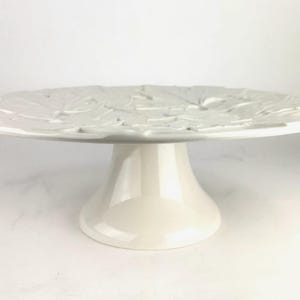 May include: A white ceramic cake stand with a raised base and a wide, round top. The top features a leaf design. The stand is approximately 10 inches in diameter and 6 inches tall, perfect for displaying cakes or desserts.