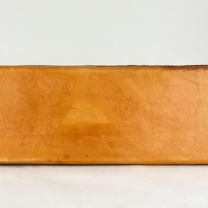 May include: A rectangular, light brown leather item with dark brown stitching along the edges. The leather has a slightly textured appearance. The item is set against a white background.