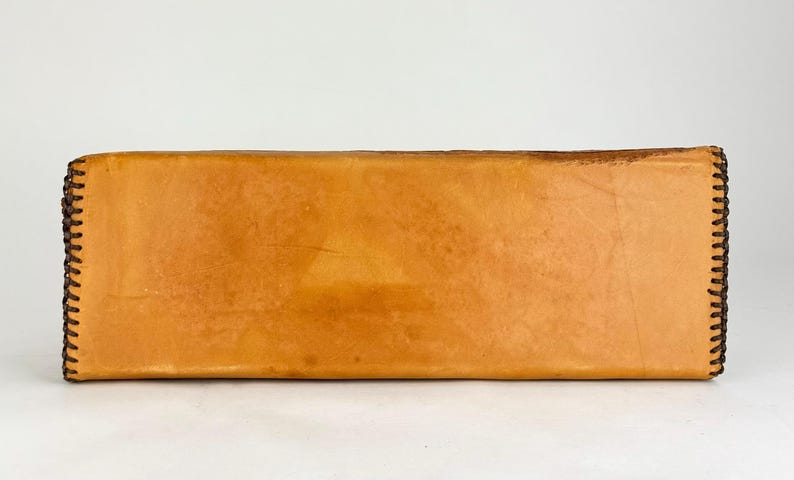 May include: A rectangular, light brown leather item with dark brown stitching along the edges. The leather has a slightly textured appearance. The item is set against a white background.