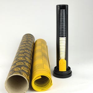 May include: A game set consisting of a cylindrical storage tube with a black and gold paisley pattern, a rolled yellow game board featuring black geometric line designs, and a vertical black plastic dispenser holding a stack of black and white discs.