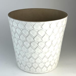 Peut inclure: Un cache-pot cylindrique en céramique présentant un extérieur blanc avec un motif en relief répétitif en forme de goutte ou d'écaille. L'intérieur du récipient est d'une couleur beige lisse et non émaillée, contrastant avec la finition blanche brillante.